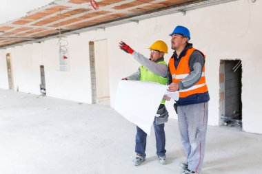İnşaat işçileri ve bir mimar ellerinde bir plan tutuyorlar ve bir inşaat sahasına bakıyorlar..