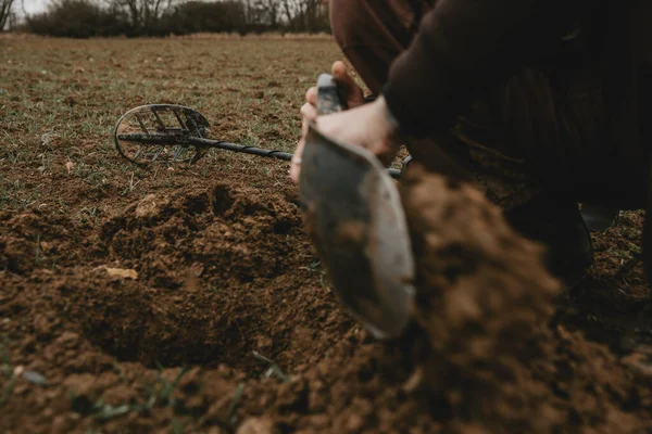 Detail of man\'s hands with shovel in it, during digging a hole in ground after positive signal of metal detector with lying metal detector in background