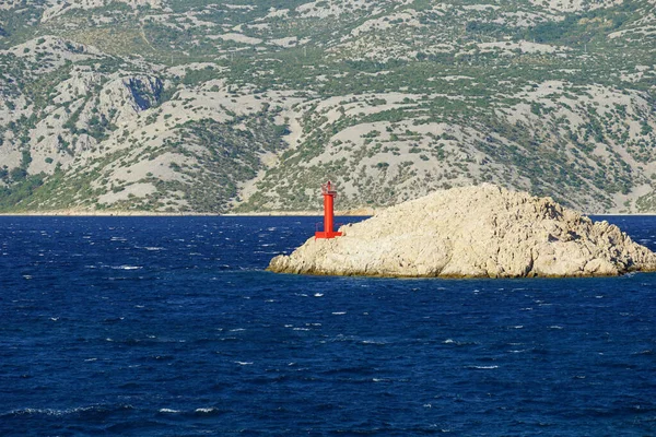 A stone island with a small red lighthouse surrounded by dark blue sea water on the sunny and windy day