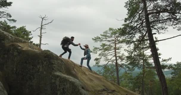 Een man helpt een vrouw een klif te beklimmen. Hij trekt aan haar hand. Ze kijken naar de natuur. Wandelen in de bergen. 4K DCI — Stockvideo