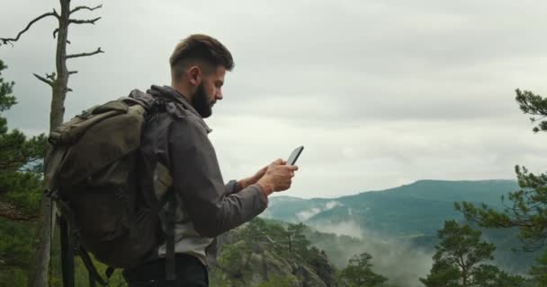 Een man staat op de top van een berg en typt een bericht op zijn smartphone. Hij heeft een grote rugzak op zijn schouders. Wandelen in de bergen. De top van de berg. 4K DCI — Stockvideo