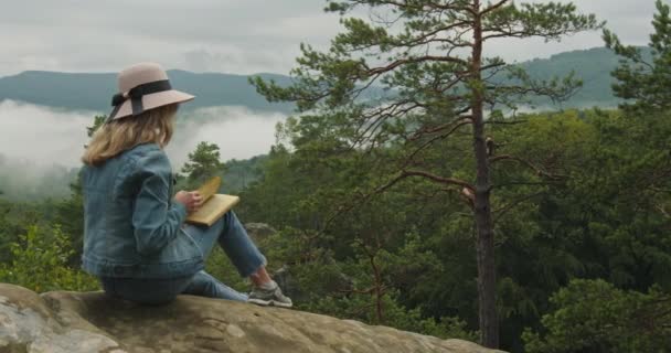 Uma mulher está sentada em cima de um penhasco e lendo um livro. Caminhadas nas montanhas. 4K DCI — Vídeo de Stock