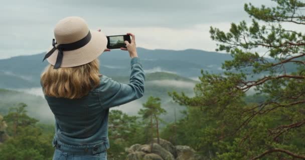 Seorang wanita berdiri di atas tebing. Dia mengambil foto pemandangan gunung yang indah. 4K DCI — Stok Video