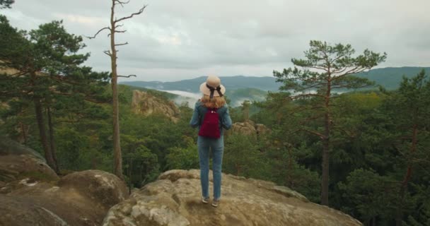 Žena stojí na vrcholu útesu. Má na ramenou červený batoh. Turistika v horách. 4K DCI — Stock video