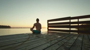 Genç bir adam iskelede oturuyor ve meditasyon yapıyor. Güzel gün batımı. Gölde dinlen. 4K