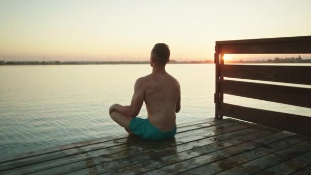 Genç bir adam iskelede oturuyor ve meditasyon yapıyor. Güzel gün batımı. 4K — Stok video