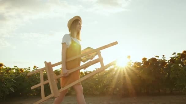 Młoda kobieta nosi sztalugę i obraz. Idzie przez pole słoneczników. Artysta w naturze. 4K — Wideo stockowe