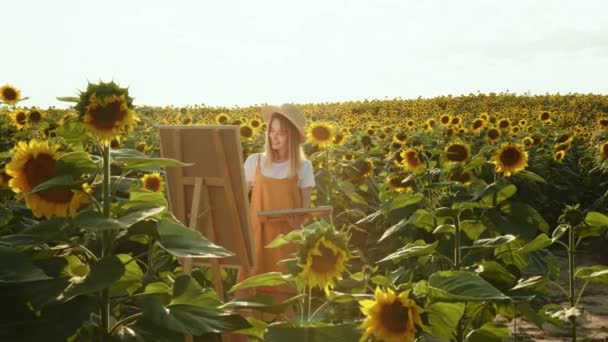 Kobieta stoi na polu słoneczników i rysuje obraz. Płótno stoi na sztaludze. Rozpuszcza kolory w palecie. Artysta w naturze. 4K — Wideo stockowe