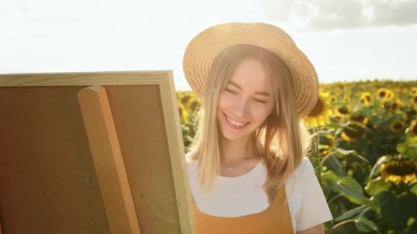 Жінка стоїть на полі соняшників і малює картину. Шеш посміхається і носить солом'яний капелюх. Художник у природі. Крупним планом стрілянина. 4-кілометровий — стокове відео