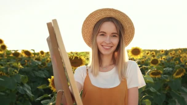 Kobieta w słomkowym kapeluszu się uśmiecha. Stoi w pobliżu sztalugi wśród słoneczników. 4K — Wideo stockowe