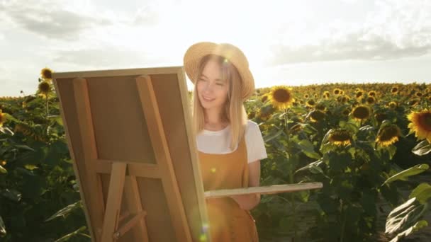 Μια γυναίκα στέκεται σε ένα χωράφι με ηλιοτρόπια και ζωγραφίζει μια εικόνα. Κοιτάζει την κάμερα και χαμογελάει. 4K — Αρχείο Βίντεο