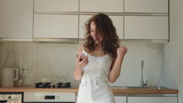 La giovane donna sorride e si rallegra. Sta ascoltando musica con le cuffie e messaggiando sul suo telefono. Lei è in cucina a casa. 4K — Video Stock