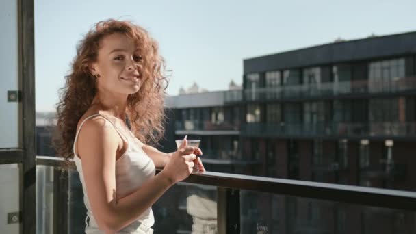 一位年轻妇女站在公寓的阳台上，端着一杯咖啡。她看着摄像机，微笑着。太阳在灿烂地照耀着.4K — 图库视频影像
