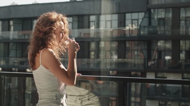 Seorang wanita muda berdiri di balkon dan minum kopi panas. Matahari bersinar dengan indah. 4K — Stok Video