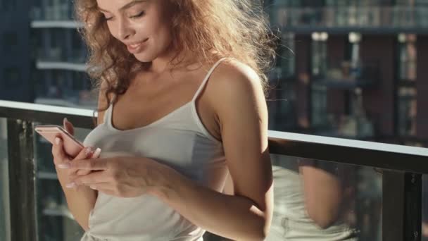 Une femme se tient debout sur le balcon et envoie des SMS sur son smartphone. Le soleil brille de mille feux. 4K — Video