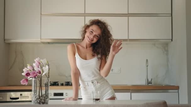 A young woman is standing in the kitchen. She is looking at the camera and waving. She is smiling. 4K Royalty Free Stock Video