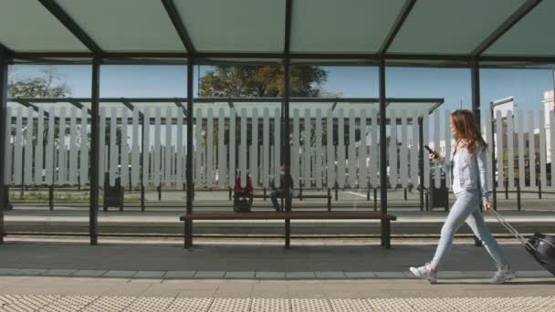 A young woman is walking with a suitcase through a public transport stop. She is texting on her smartphone. 4K — 图库视频影像