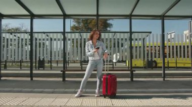 A young woman is standing at a public transport stop. She smiles. She is holding a suitcase in her hand and texting on her smartphone. 4K