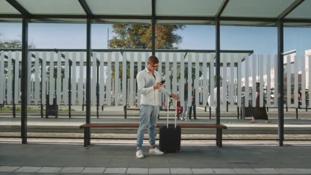 En mand står ved et stoppested. Han holder en kuffert i hånden og sms 'er på sin smartphone. 4K – Stock-video