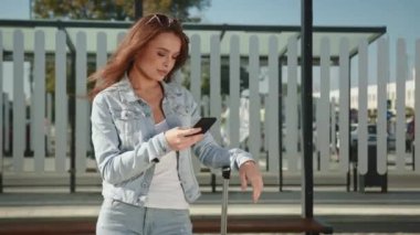 Genç bir kadın toplu taşıma durağında duruyor. Elinde bir bavul tutuyor ve akıllı telefonuyla mesaj atıyor. Rüzgar saçlarını savuruyor. 4K