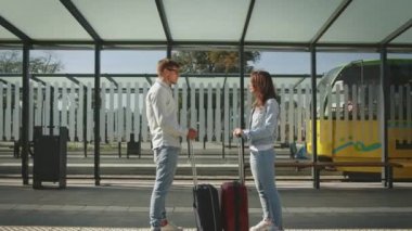 Bir adam ve bir kadın toplu taşıma durağında duruyorlar. Duygusal konuşuyorlar ve bavullarını tutuyorlar. Arka planda bir tramvay var. 4K