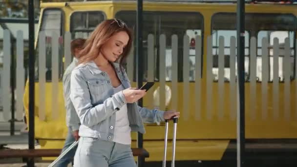 En ung kvinde står ved et stoppested. Hun holder en kuffert i hånden og sms 'er på sin smartphone. En gammel sporvogn kører i baggrunden. 4K – Stock-video