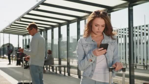 En ung kvinde sms 'er på sin smartphone. Hun står ved et stoppested for offentlig transport. Hun har en kuffert. Folk går i baggrunden. 4K – Stock-video
