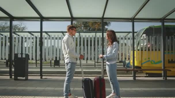 男と女が公共交通機関の停留所に立っている。彼らは感情的に話し、スーツケースを持っています。路面電車が運転している。4K — ストック動画