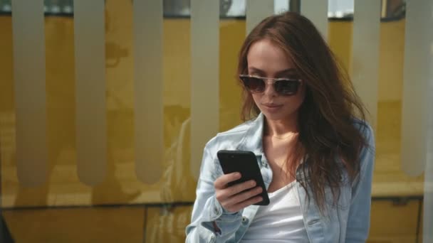 Eine junge Frau sitzt an einer Bushaltestelle und schreibt SMS auf ihr Smartphone. Im Hintergrund fährt eine alte Straßenbahn. 4K — Stockvideo