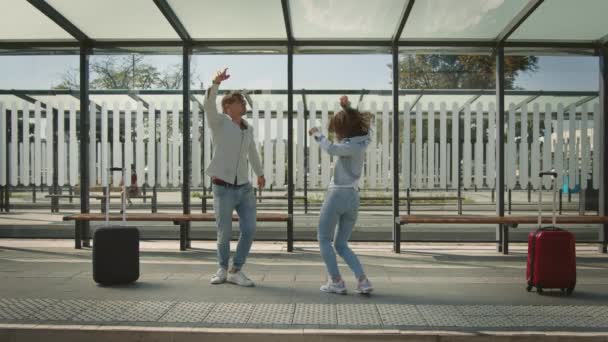 Un uomo e una donna ballano emotivamente alla fermata dell'autobus. Si stanno divertendo e ridendo. Ci sono delle valigie accanto a loro. 4K — Video Stock