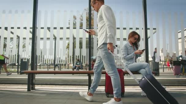 Une jeune femme est assise à un arrêt de transport en commun et envoie des SMS sur son smartphone. Un homme est assis à côté d'elle et envoie aussi des textos. 4K — Video