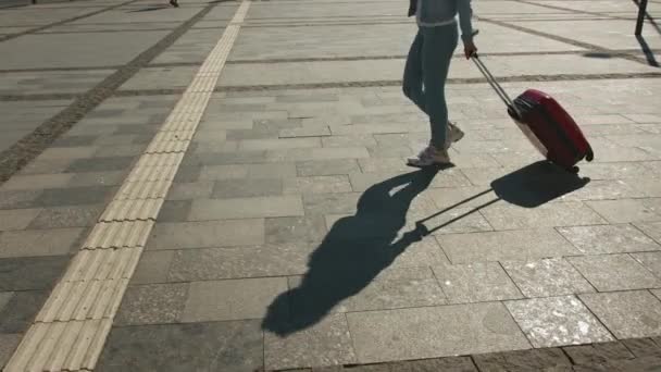 Una giovane donna sta camminando attraverso la stazione ferroviaria. Sta arrotolando una valigia. Potete vedere la sua ombra sul marciapiede. 4K — Video Stock