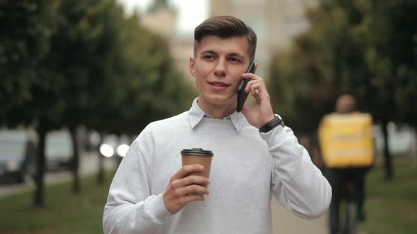 Seorang pria berbicara secara emosional di telepon. Dia minum kopi dan berjalan melewati taman. 4K — Stok Video