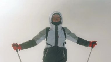 Genç bir kadın bir dağın tepesinde duruyor ve kameraya bakıyor. Elinde trekking sopaları tutuyor. Güçlü rüzgar ve kar var. Pikui Dağı. 4K