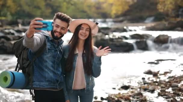 Una giovane donna e un uomo sono sulla riva di un fiume di montagna. Si stanno facendo dei selfie. Sorridono e salutano le mani. Escursioni in montagna. 4K — Video Stock