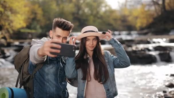 Uma jovem e um homem estão à beira de um rio de montanha. A mulher está a ajustar o chapéu. Eles estão tirando selfies e sorrindo. 4K — Vídeo de Stock