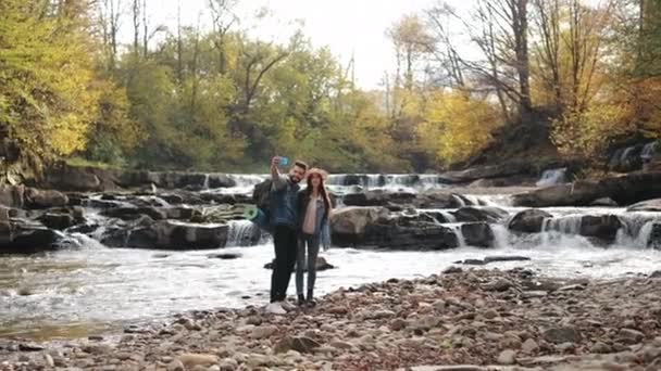Une jeune femme et un homme se tiennent sur le rivage d'une rivière de montagne. Ils prennent des selfies et sourient. Randonnée dans les montagnes. 4K — Video