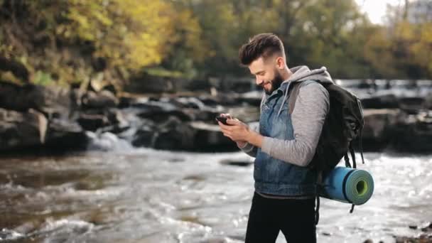 Un hombre está de pie en la orilla de un río de montaña y mensajes de texto en un teléfono inteligente. Senderismo en las montañas. 4K — Vídeo de stock
