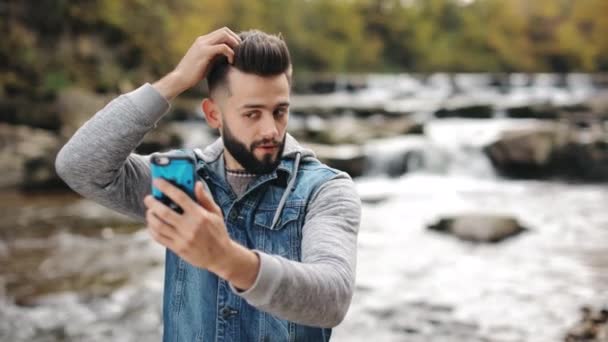 Bir adam selfie çekiyor ve saçını düzeltiyor. Bir dağ nehrinin arka planında duruyor. Dağlarda yürüyüş. 4K — Stok video