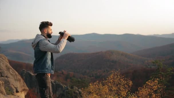 그 남자는 벼랑 끝에서 서 쌍안경으로 바라본다. 뒤에 산 풍경이 있습니다. 산에서 하이킹도 하고. 4K — 비디오