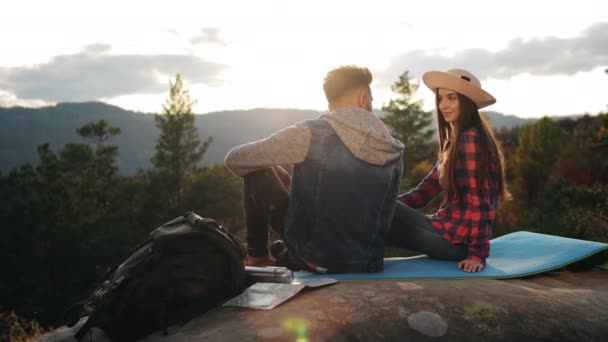 Uma jovem e um homem estão sentados numa grande rocha. Estão a olhar um para o outro. Pôr-do-sol, montanha. 4K — Vídeo de Stock