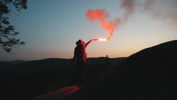 Une jeune femme est debout sur le bord d'une falaise et agite des fusées éclairantes dans la nuit. Randonnée dans les montagnes. 4K — Video