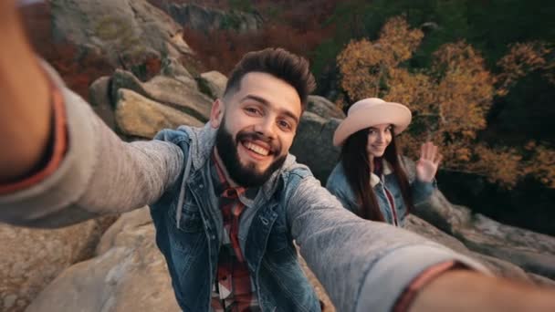 A young man is shooting a video on the front camera. A smiling woman is standing near his and waving her hand. Mountain landscape in the background. 4K — Stock Video