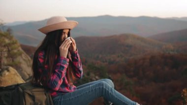 Genç bir kadın sıcak çay içiyor. O bir kayanın üzerinde oturuyor ve dağ manzarasına bakıyor. Dağlarda yürüyüş. 4K