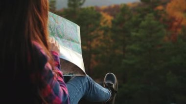 Genç bir kadın bir kayanın üzerinde oturuyor ve elinde bir harita tutuyor. Bir rota çiziyor. Dağlarda yürüyüş. 4K