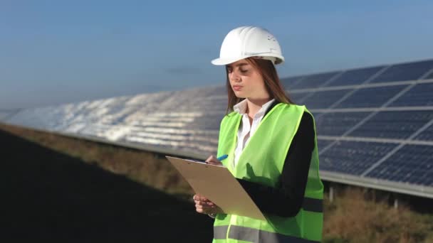 Инженер-женщина проводит осмотр солнечных панелей. Она вводит данные в документ. На ней зеленый жилет и каска. Солнечные панели на заднем плане. 4k — стоковое видео