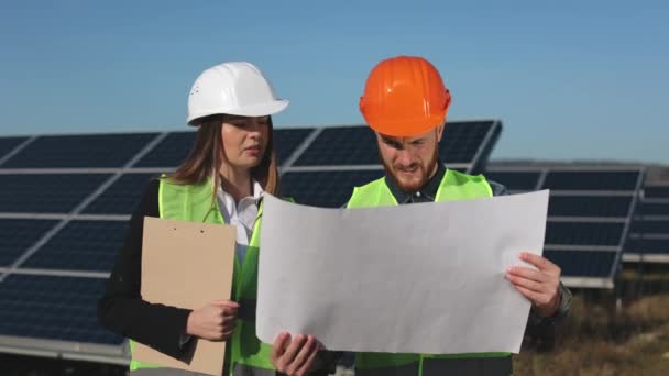 Два инженера обсуждают чертежи для проекта. Они стоят на солнечной станции. Женщина-инженер указывает рукой на объект. 4K — стоковое видео