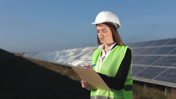 A female engineer is conducting an inspection of solar panels. She is entering data into the document. Solar panels in the background. 4k — Stock Video