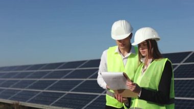 İki mühendis belgeleri kontrol edip tartışıyor. Güneş panellerinin yanında duruyorlar. 4K