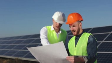 İki mühendis proje planlarına bakıyorlar. Tüm aşamaları tartışıyorlar. Arka planda güneş panelleri var. 4K